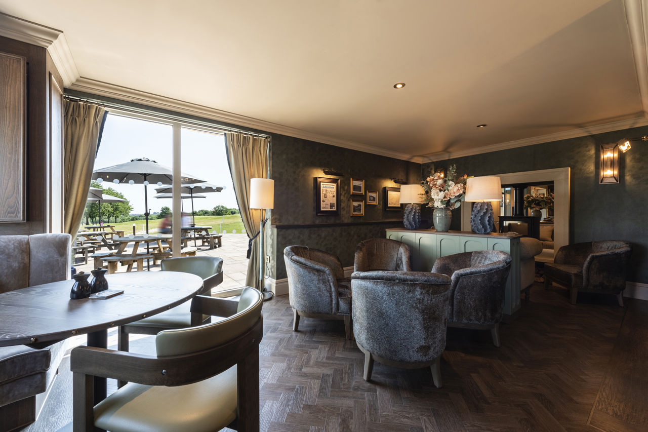 A photo of the snug area of Cook House Bar & Kitchen. A dining table and chairs are on the left of the image and a group of four armchairs are towards the centre in front of a console table holding a large vase of flowers and two lamps. A double sided stove can be seen beyond the table. Picnic benches with parasols can be seen through the large full length windows.